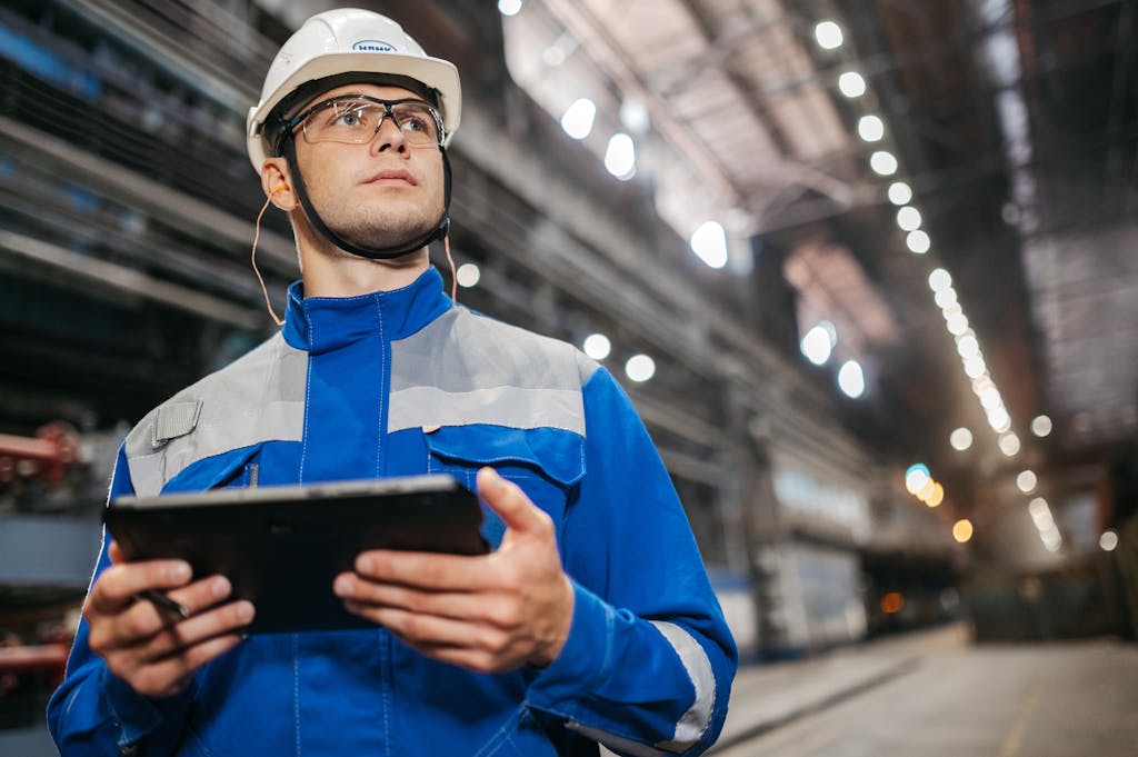 Factory worker in safety gear using a tablet in a well-lit industrial setting.
